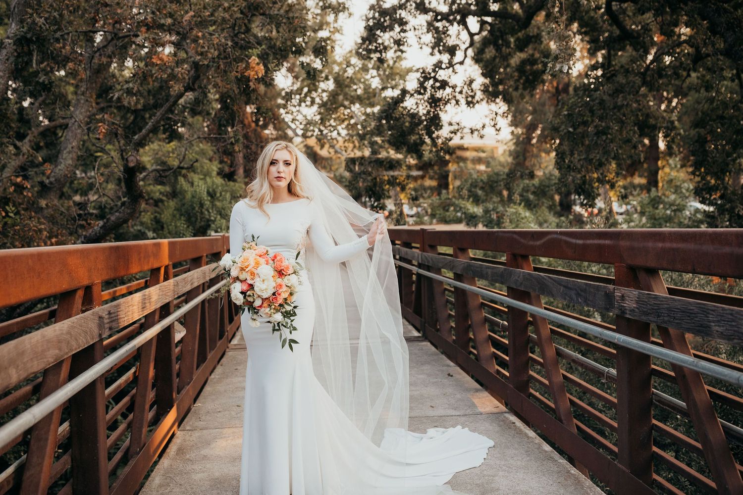 Bride in white lace gown with flowing veil stands on wooden bridge holding orange and pink bouquet at sunset.