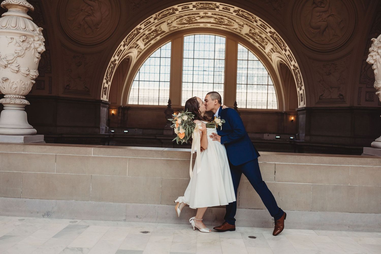 A couple in formal wedding attire dances and poses together in an elegant historic hall with ornate architecture.