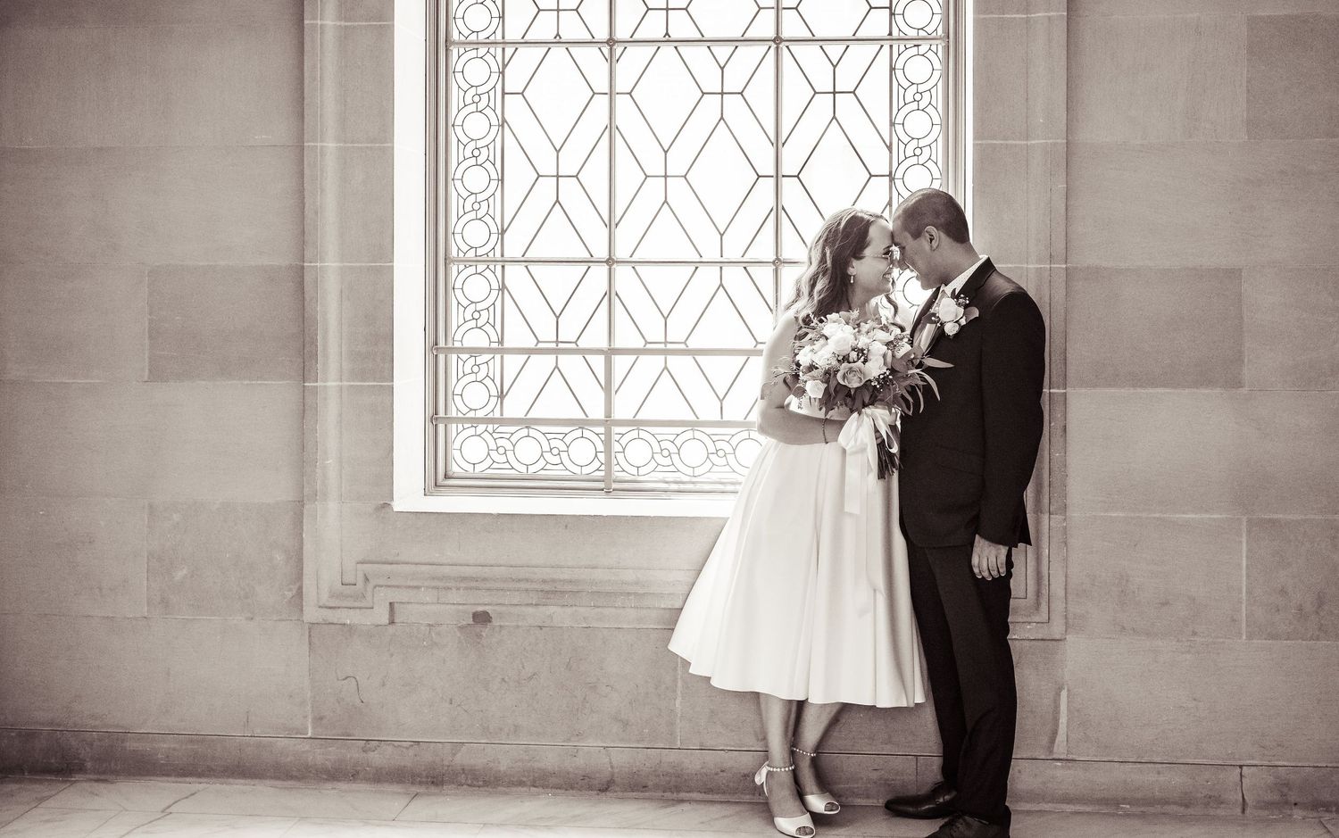 A romantic black and white wedding photo of a couple embracing by an ornate window in a vintage-style setting.