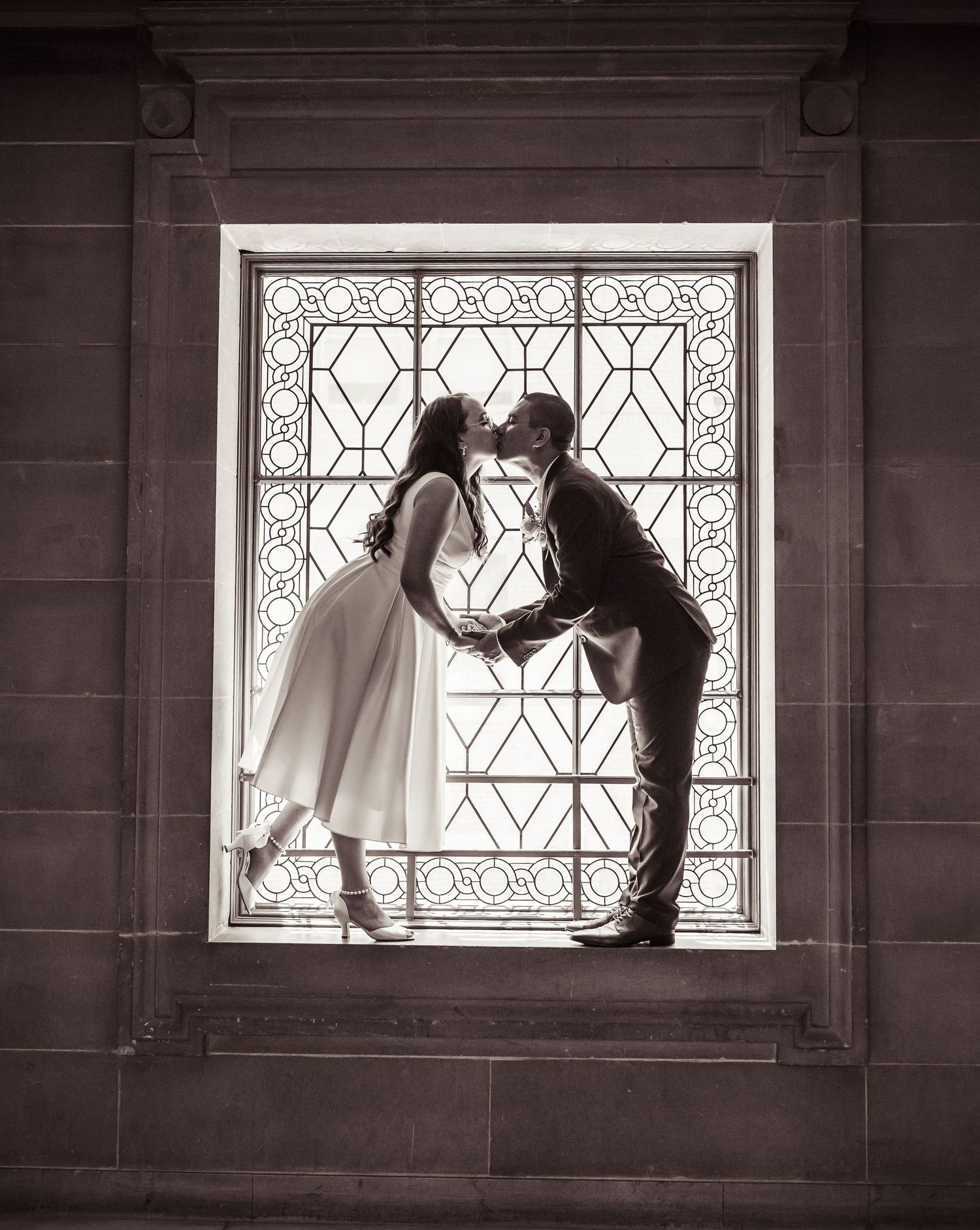 A romantic black and white silhouette of a couple kissing in front of an ornate window with decorative ironwork design.
