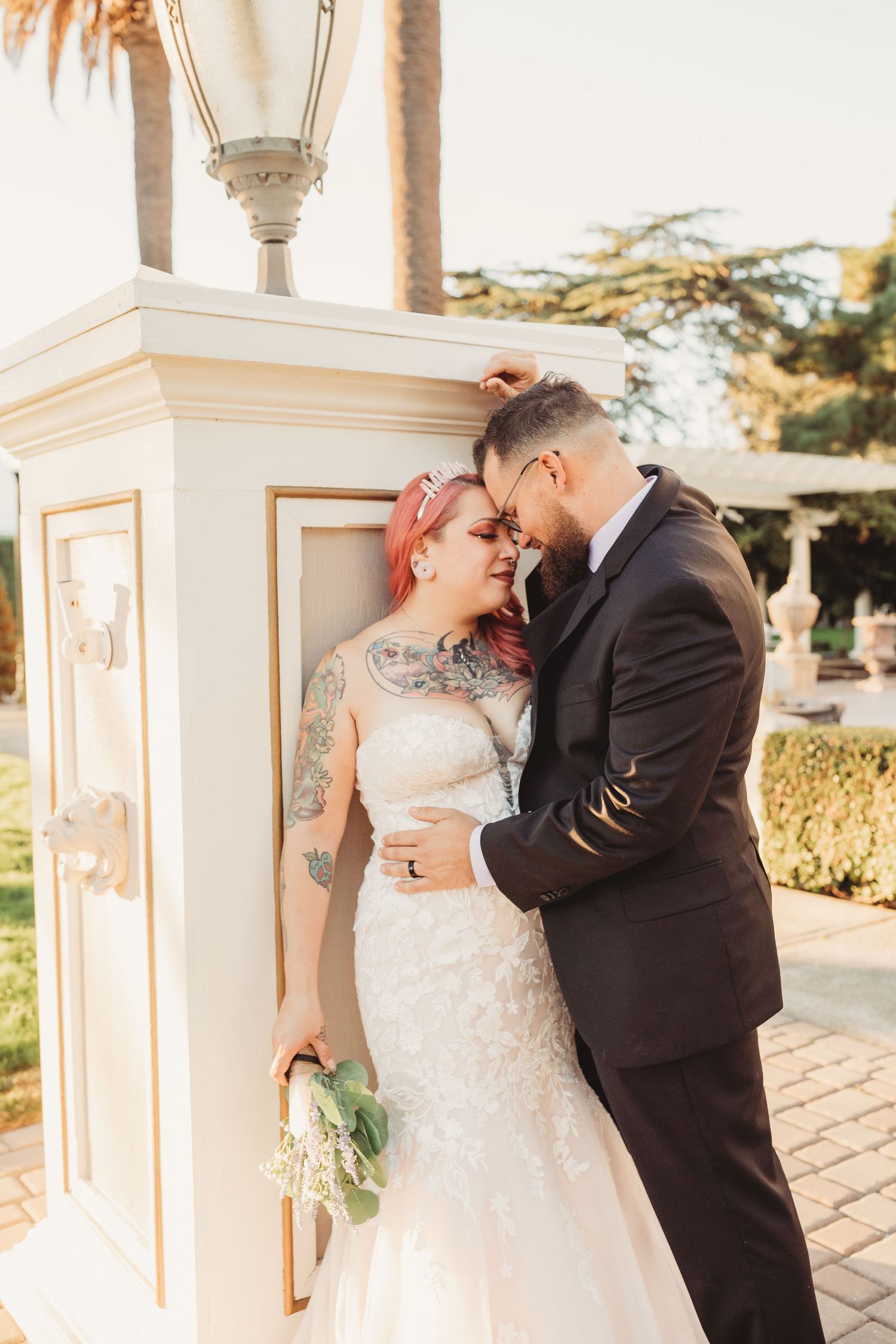 A romantic wedding couple embrace and kiss near a decorative white pillar in warm evening light.
