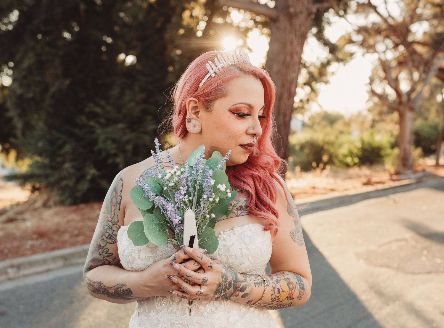 Woman with pink hair and tattoos holds spring flowers in a nature setting during golden hour.