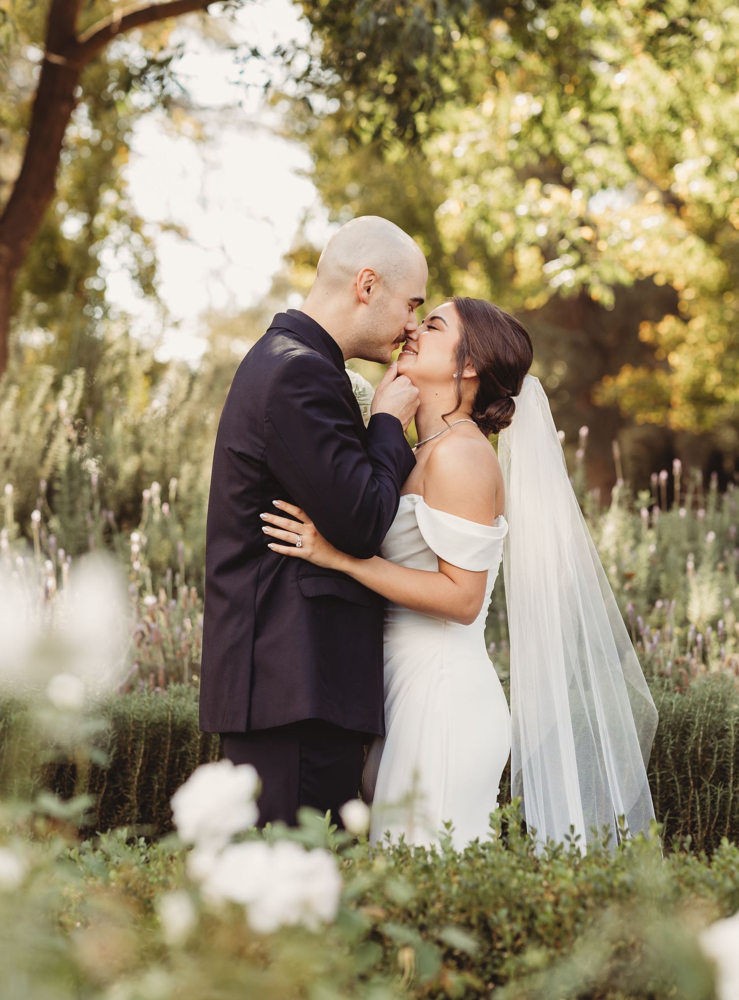 A romantic wedding couple shares a tender kiss among blooming lavender and trees in a dreamy garden setting.