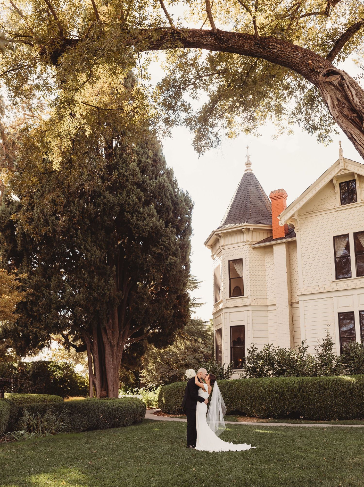A couple embraces on the lawn of a grand Victorian mansion with turret and white exterior on a golden autumn evening.