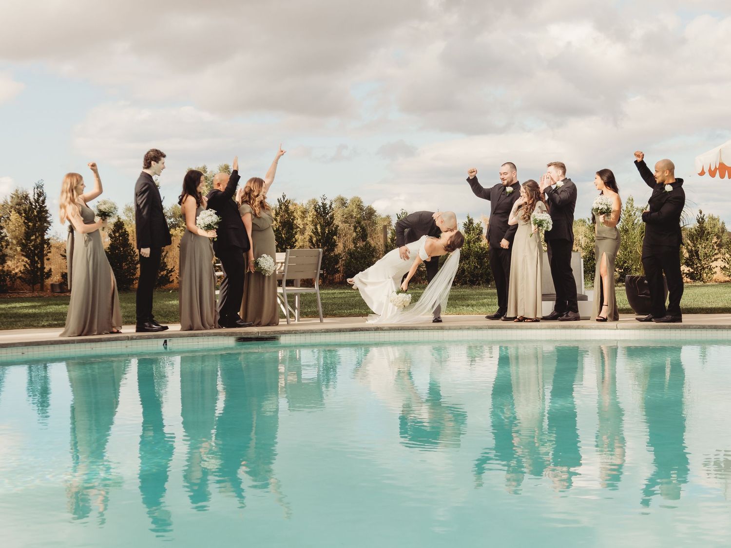 Wedding party celebrates with joyful poses around a resort swimming pool on a sunny day.
