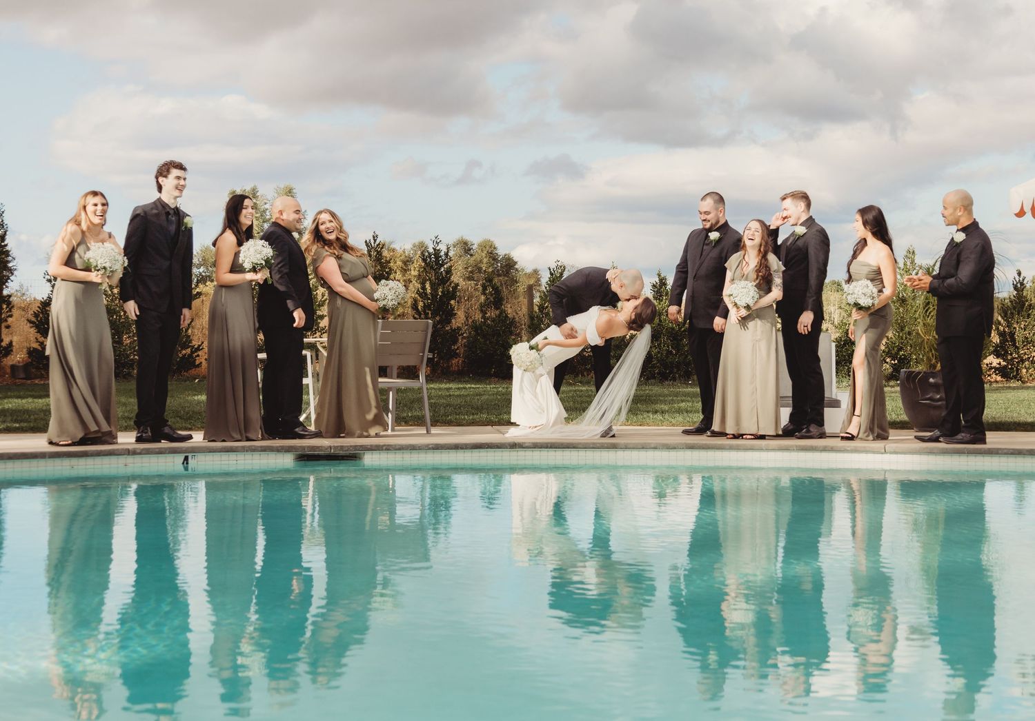 A wedding party stands along the edge of a swimming pool in formalwear during golden hour.