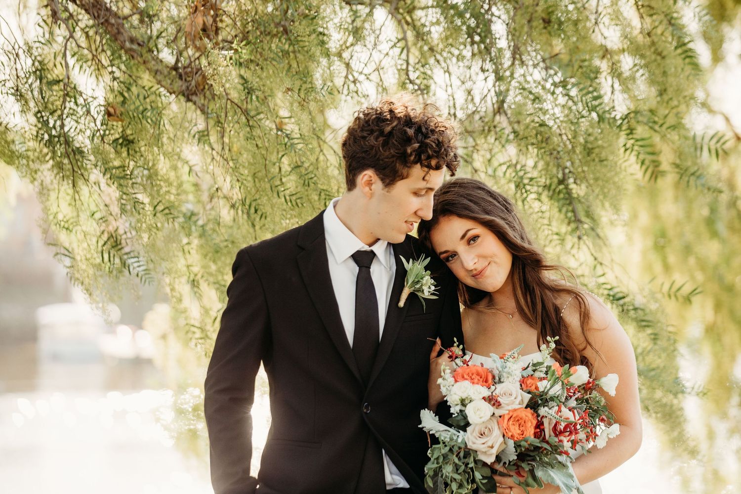 Newlywed couple sharing intimate moments under willow trees with peach and coral floral bouquet details.