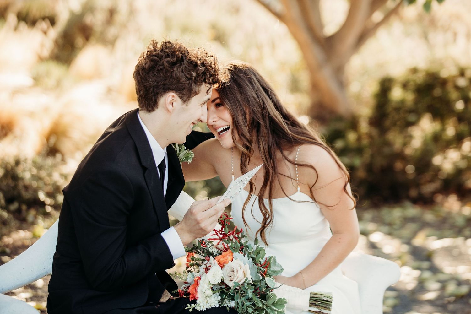 A newlywed couple shares an intimate moment in a sunlit garden setting with a bouquet of flowers.