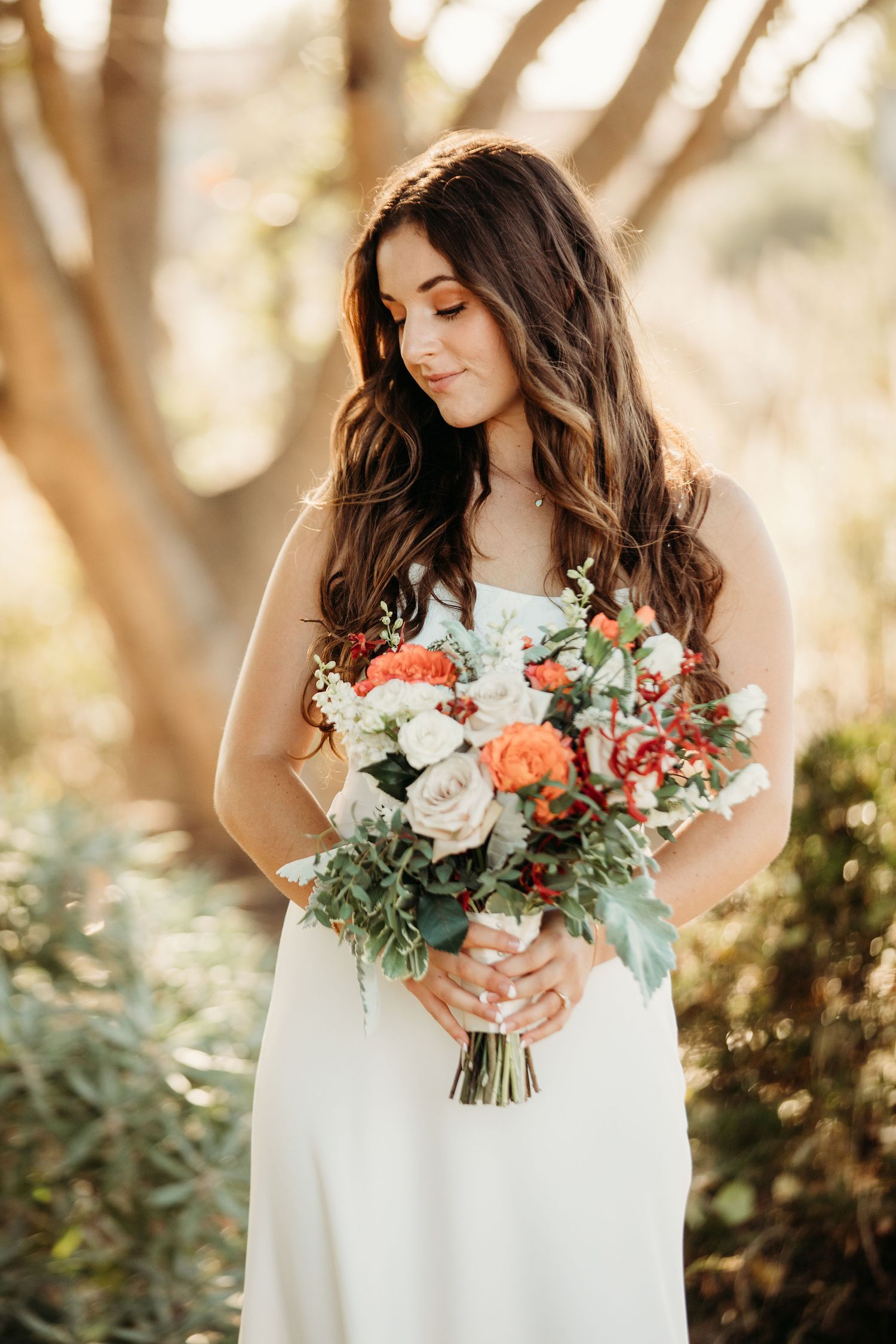 A bride in a white wedding dress holds a beautiful bouquet of coral, white and green flowers in natural outdoor lighting.