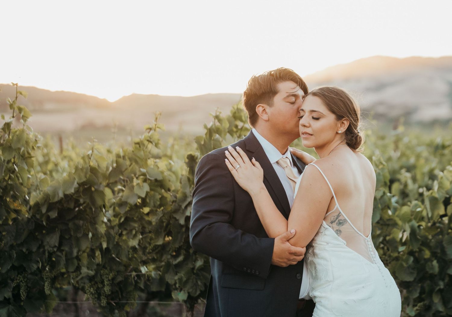 Romantic couple in wedding attire share tender kisses and embraces in a beautiful vineyard at sunset.