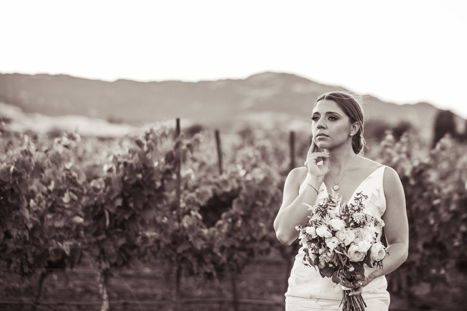 A bride poses thoughtfully in a vineyard while holding a beautiful floral bouquet in black and white.