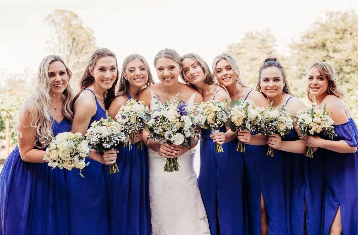A wedding party wearing deep royal blue bridesmaid dresses holding white and green floral bouquets poses outdoors.