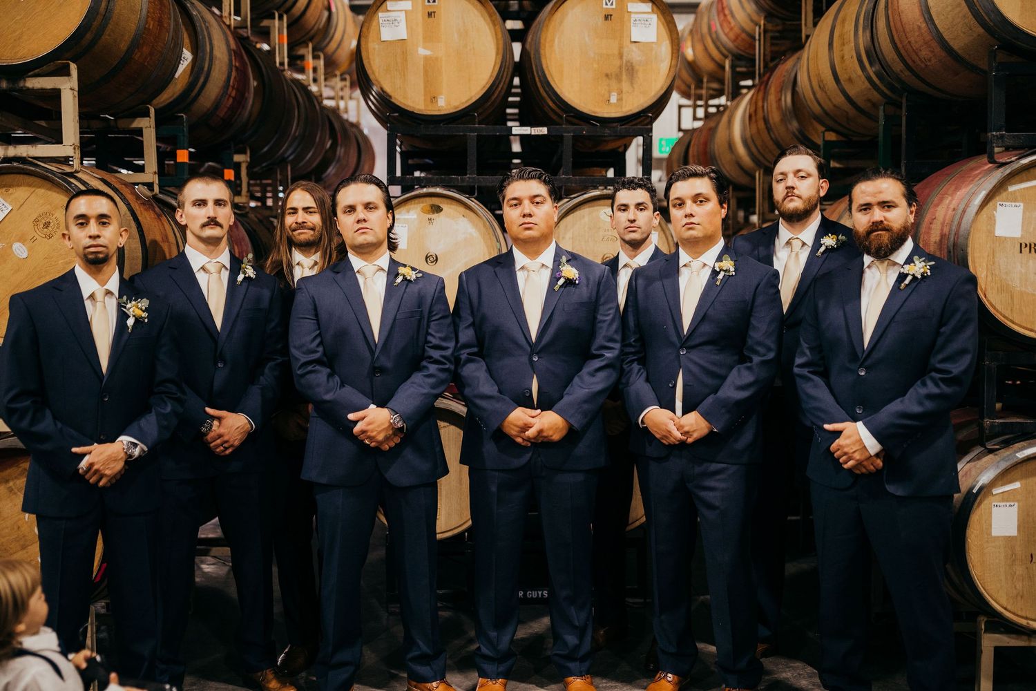 Groomsmen in navy blue suits pose together in front of wooden wine barrels at a winery venue.