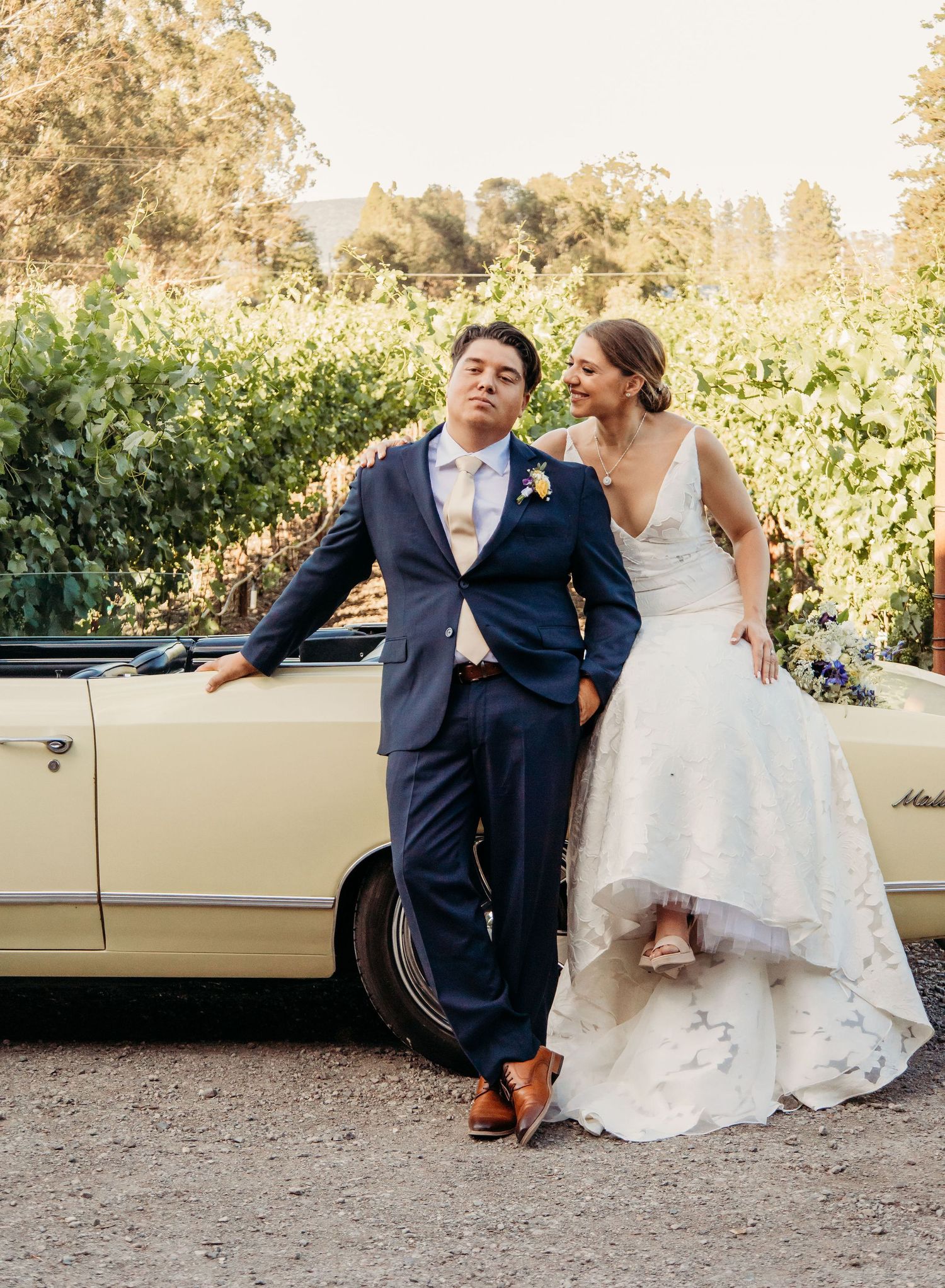 Newlyweds pose together against a vintage cream-colored car in a vineyard setting during golden hour.