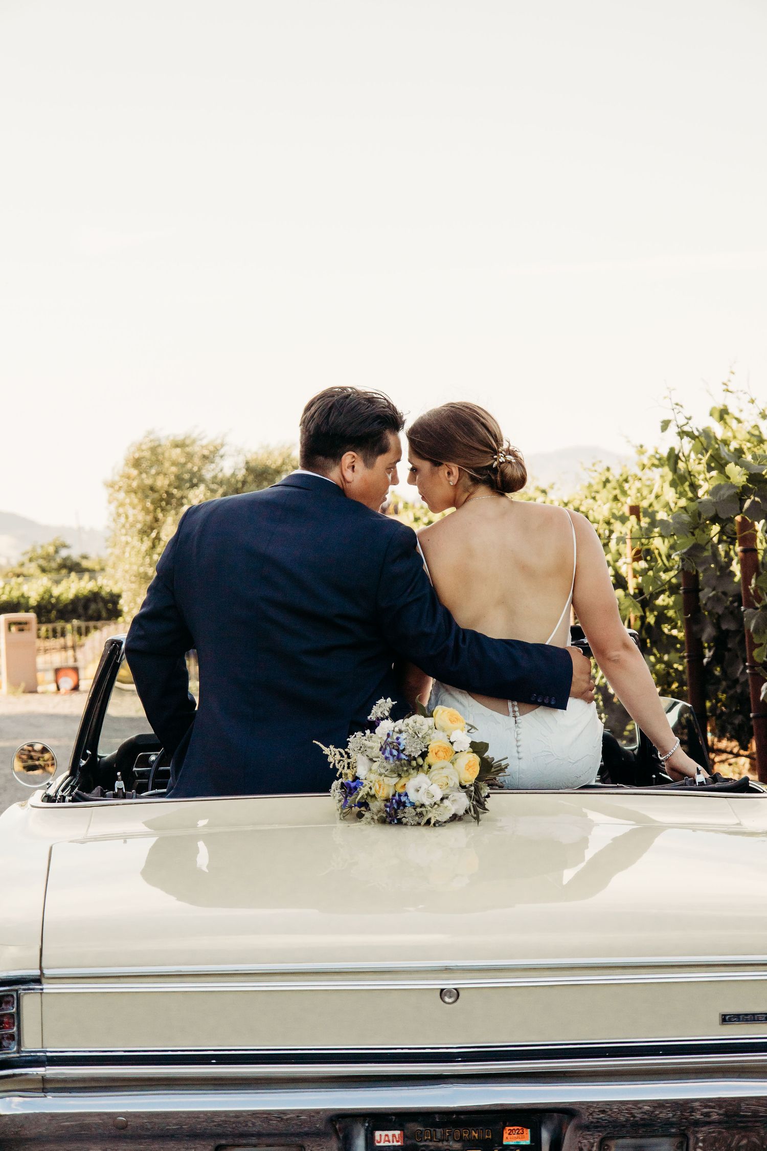 Newlywed couple sitting on vintage car trunk with bouquet at sunset in vineyard setting.
