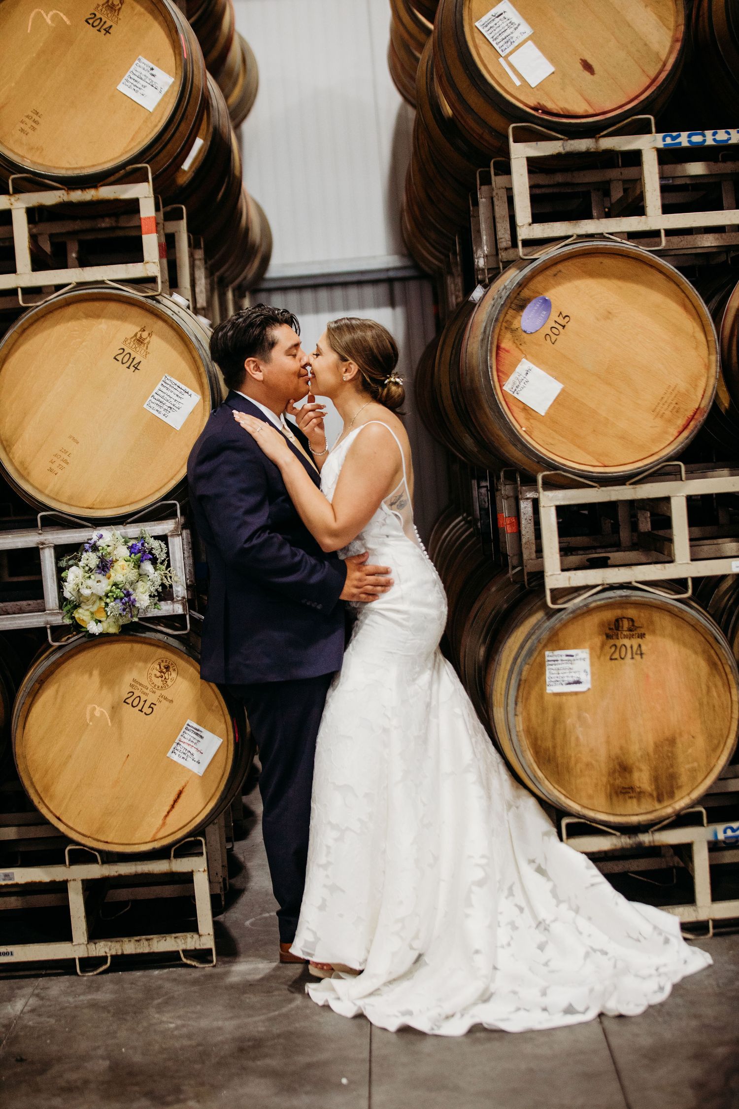 Couple in wedding attire share a romantic moment among stacked wooden wine barrels in a winery cellar.