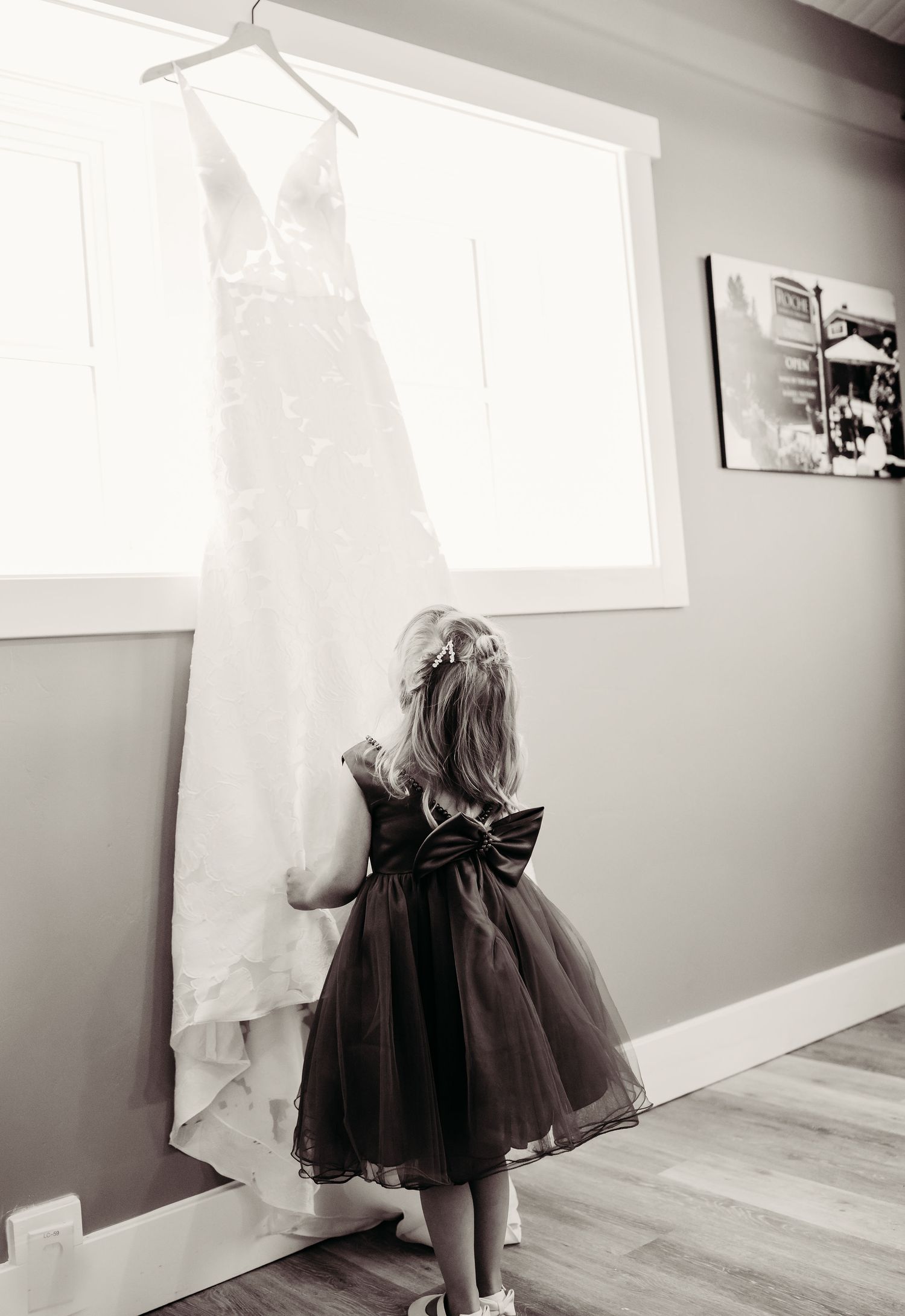 A little girl in a black tulle dress admires a white wedding gown hanging by a window in black and white photography.