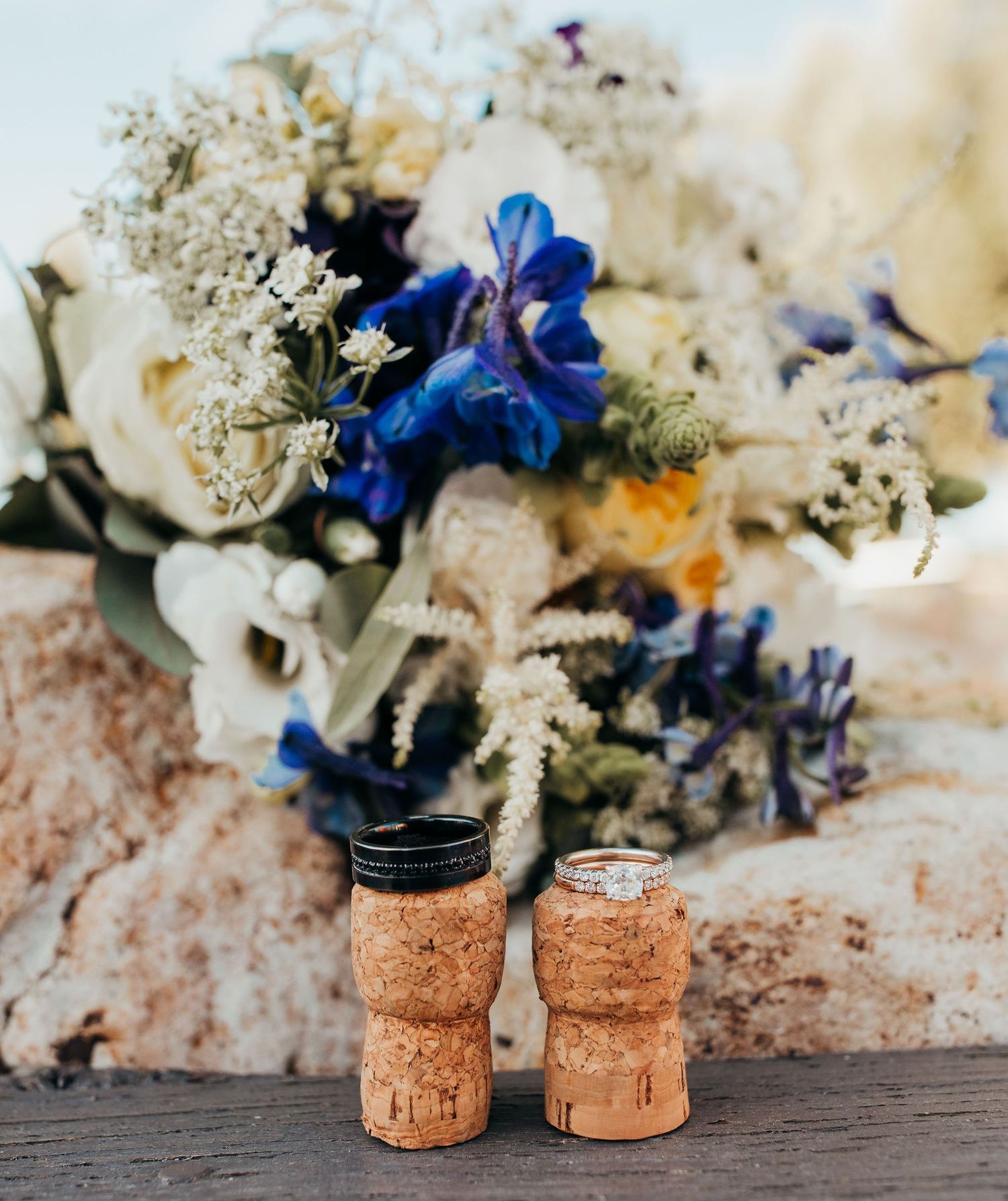 Wedding rings sit on decorative cork stoppers with a bouquet of white and blue flowers in the background.