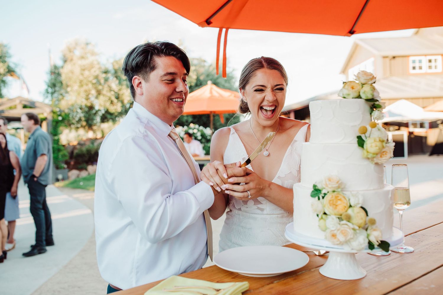 A joyful couple in wedding attire shares a laugh while cutting their white and yellow floral decorated cake outdoors.