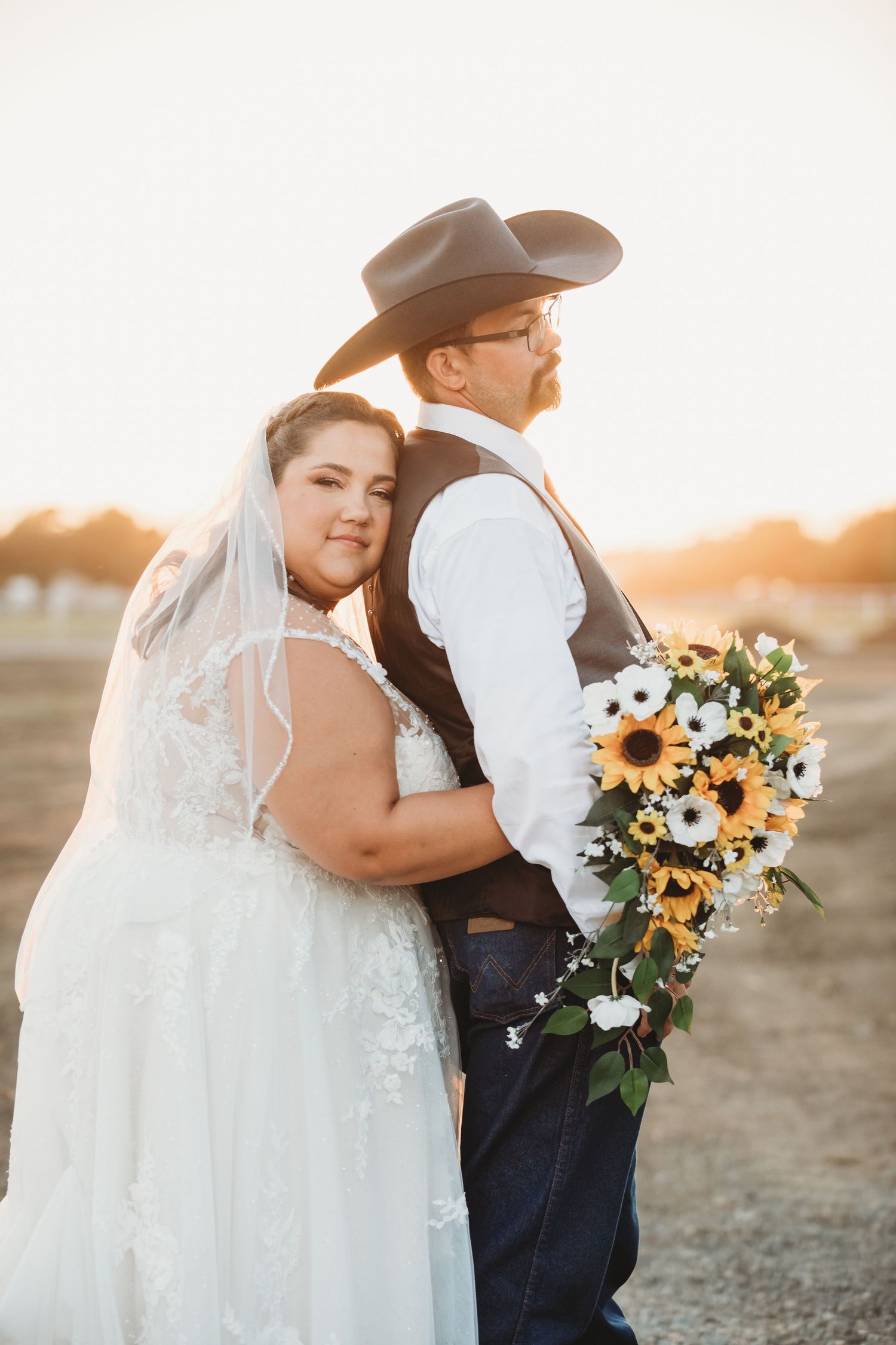 A couple embraces during their sunset wedding ceremony, with a bouquet of sunflowers and white flowers.