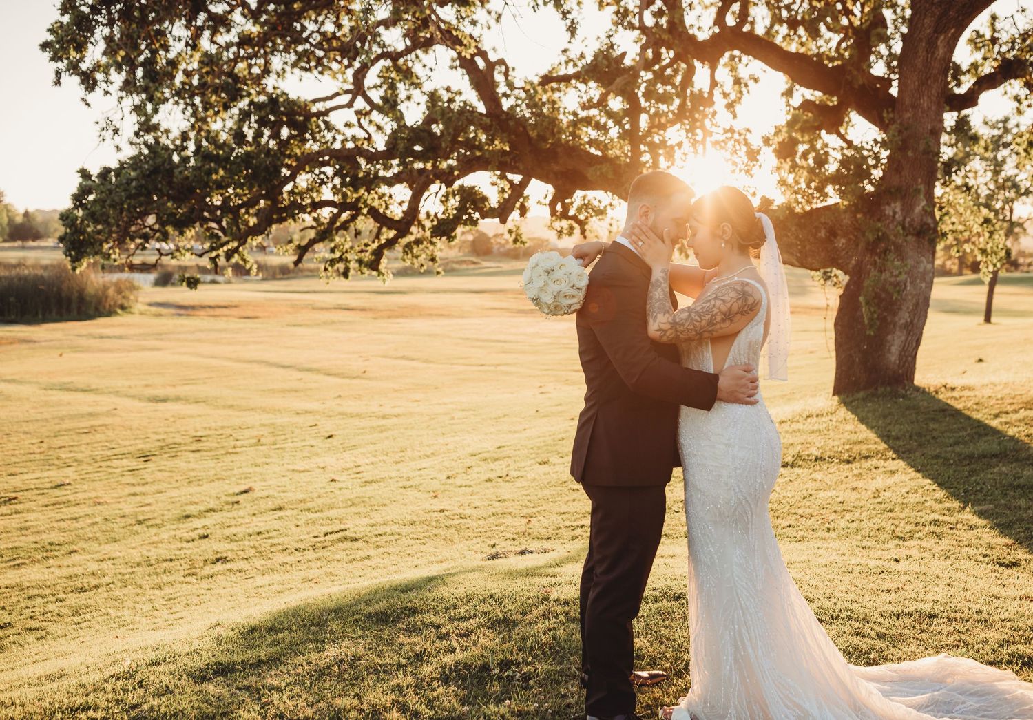 A couple shares a romantic moment at sunset under a large oak tree on a golden grass field.