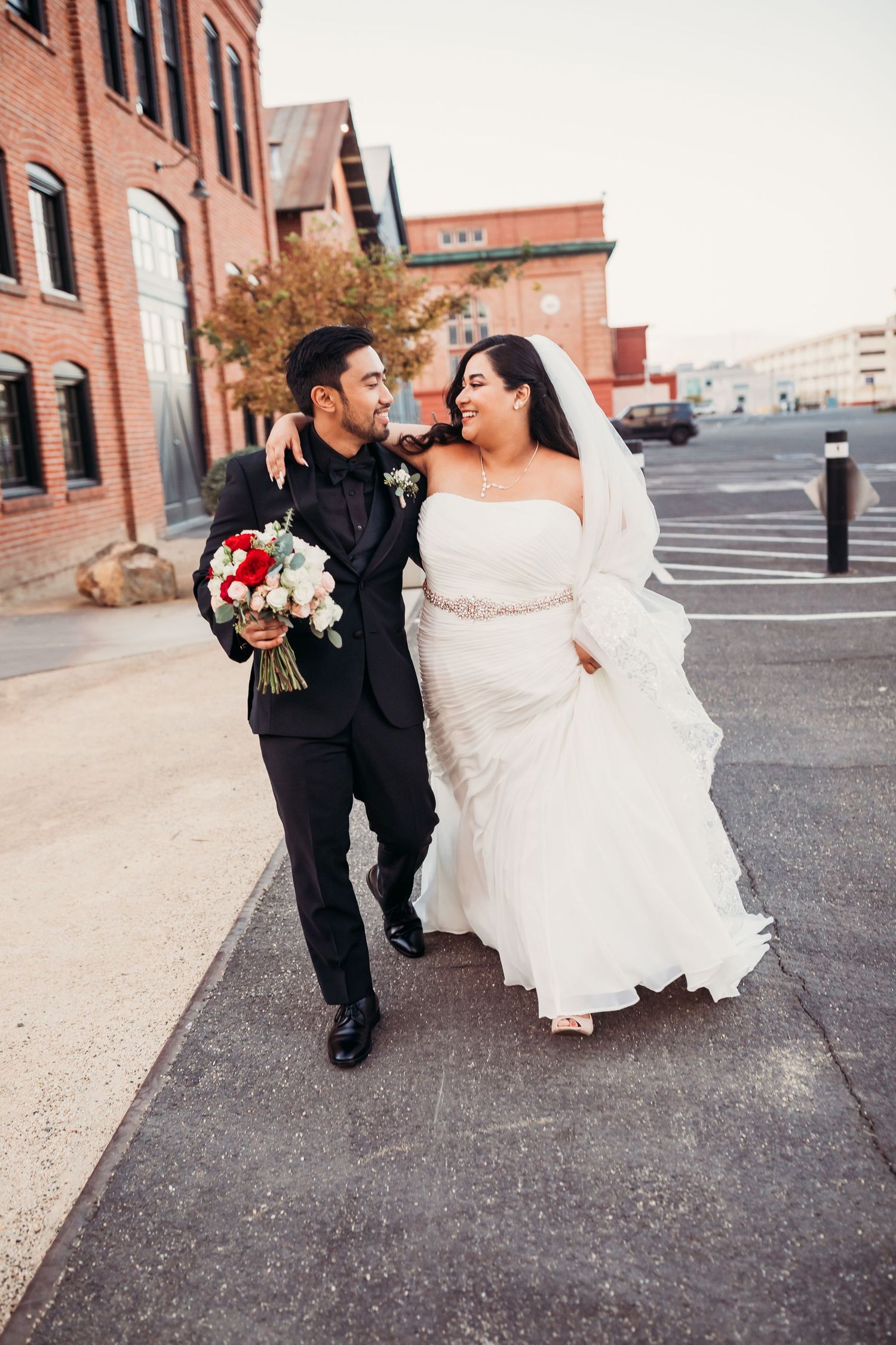 A couple in wedding attire shares a joyful moment while walking together down a brick-lined urban street.