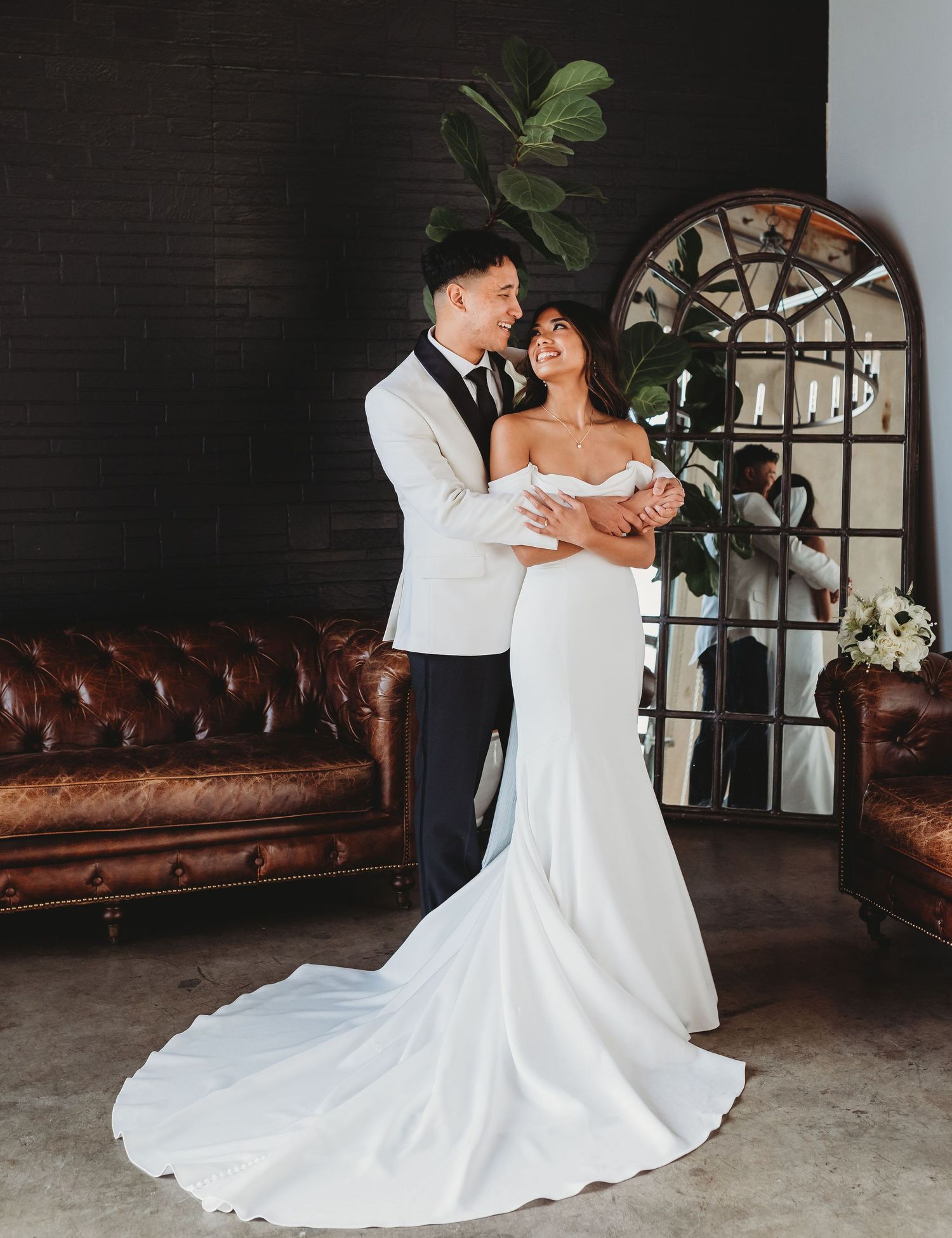 Elegant couple poses by brown leather couch and arched mirror in romantic wedding photoshoot.