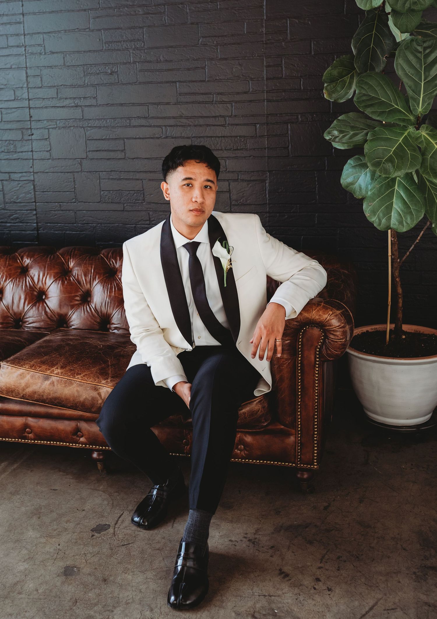 A stylishly dressed person in a white blazer and black tie sits on a brown leather Chesterfield sofa next to a potted plant.