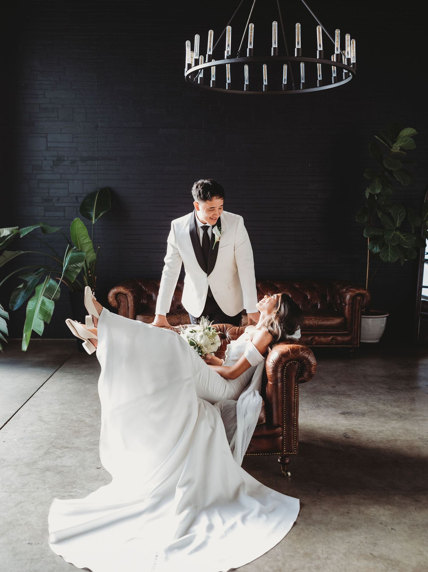 A bride in a white gown reclines on a leather sofa while someone in a white tuxedo jacket stands beside her under a chandelier.