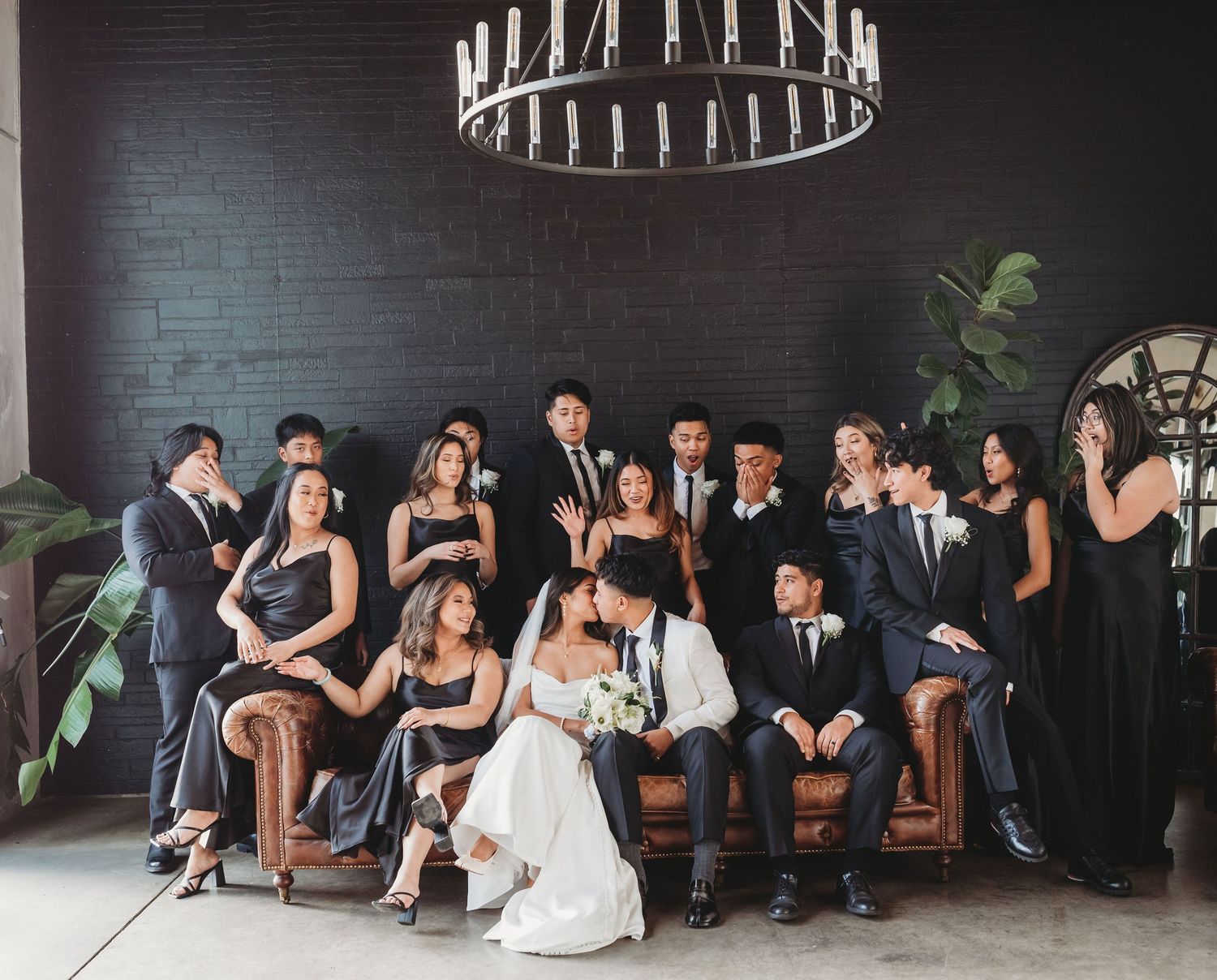 Wedding party poses elegantly in formal black attire against dark brick wall with modern circular chandelier above.