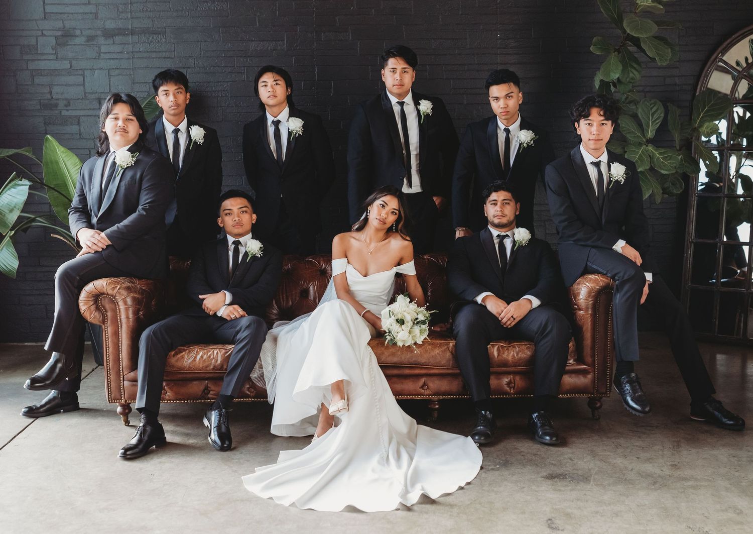 Wedding party in formal attire gathered elegantly around a brown leather couch against textured grey brick wall.