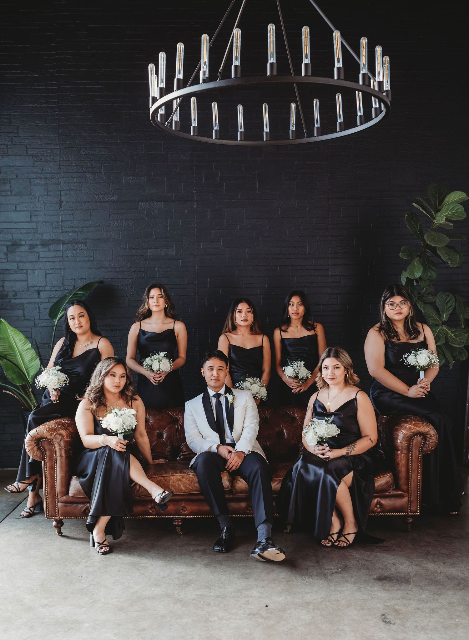 A bridal party lounges on a brown leather couch against a dark wall with modern lighting fixtures above.