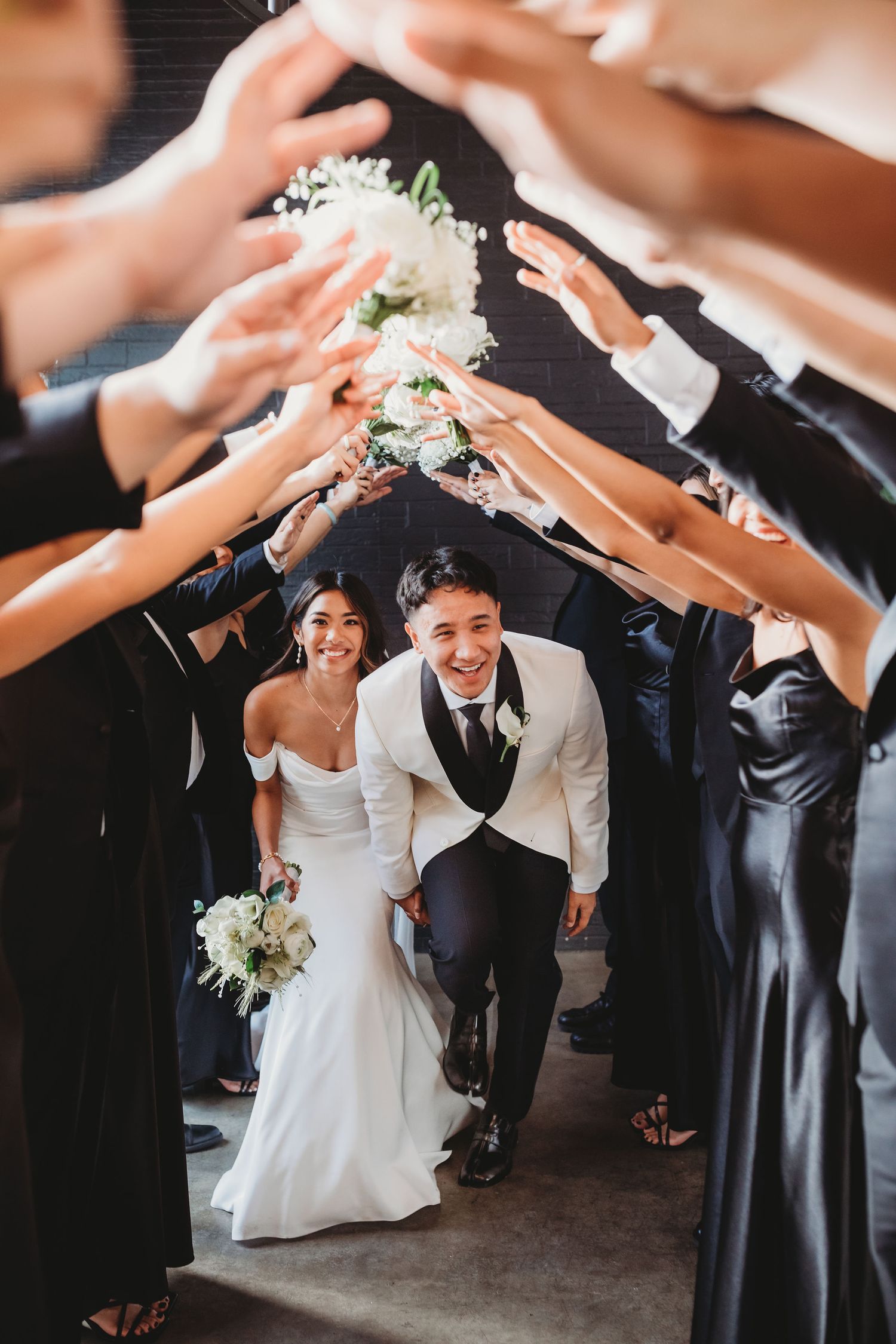 Newlyweds joyfully walk through a celebratory tunnel of raised hands made by wedding guests at an indoor venue.