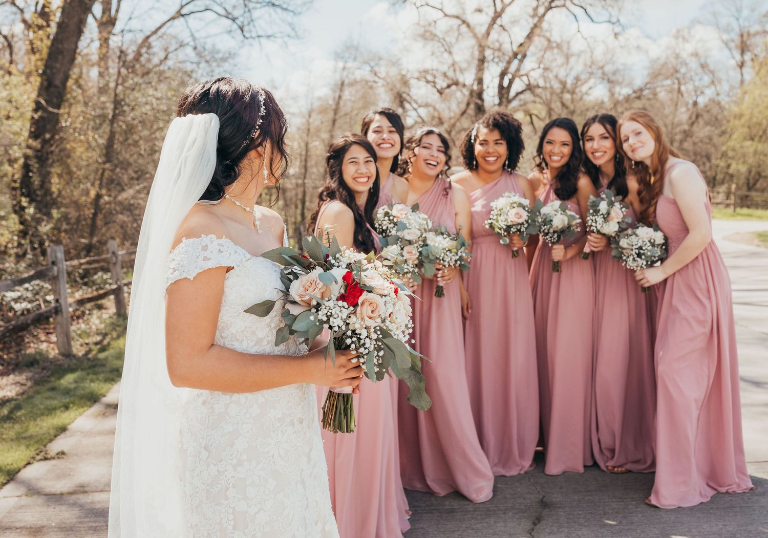 A wedding party gathers outdoors wearing white and dusty rose gowns while holding floral bouquets.