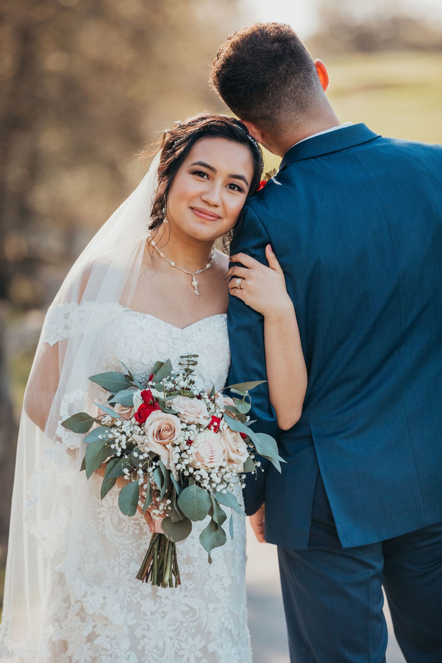 Wedding couple poses outside with lush bouquet featuring pink roses and eucalyptus greenery against a soft background.