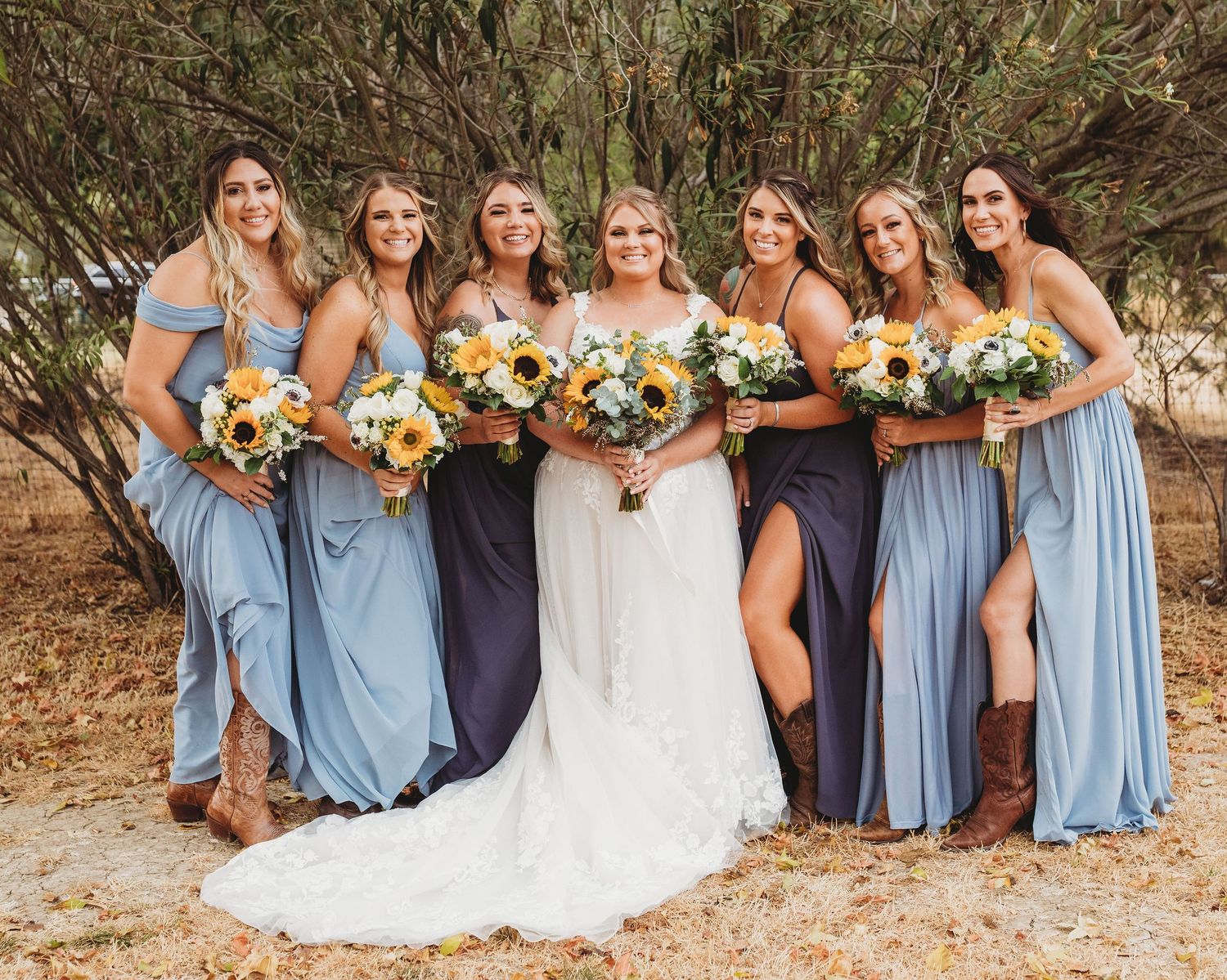 A group in powder blue bridesmaid dresses and a white wedding gown pose together holding sunflower bouquets outdoors.