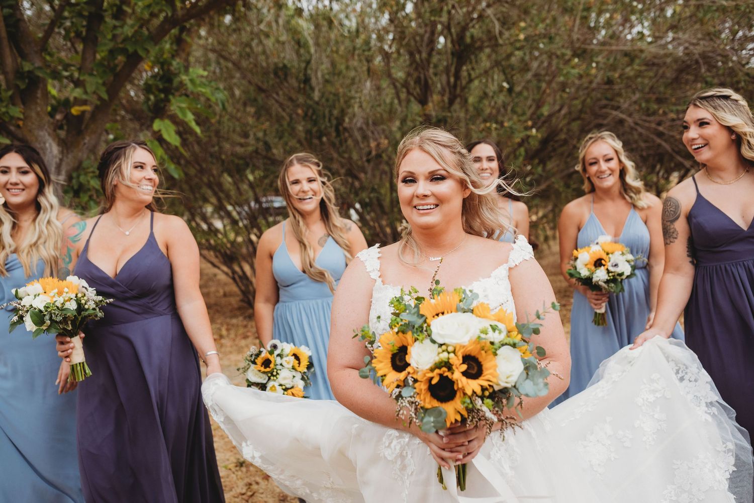 Bridal party in blue dresses poses outdoors with bright sunflower bouquets during rustic garden wedding.