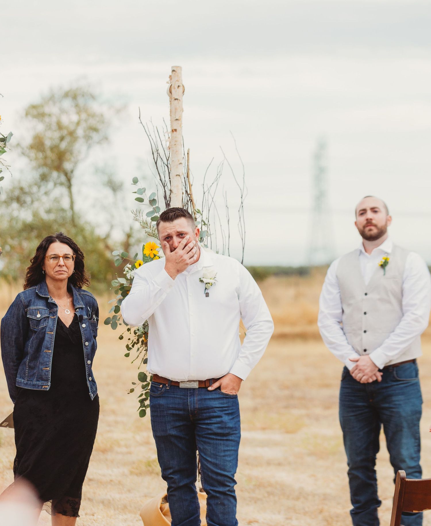 Three people stand together at an outdoor rustic wedding ceremony site with bare branches and floral decor.