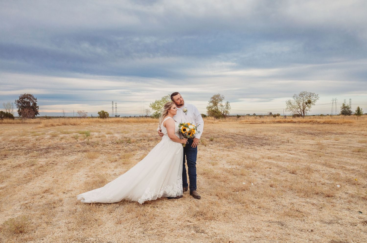 A couple in wedding attire embrace in a golden Australian outback setting with cloudy skies and sparse vegetation.