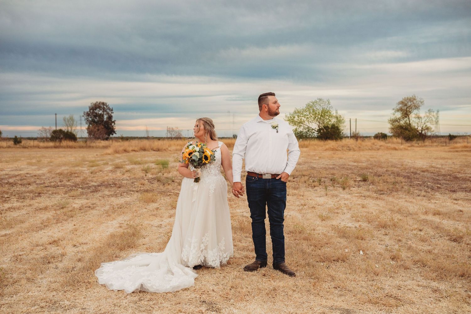 A couple stands together holding hands in a rustic field at sunset with cloudy skies overhead.