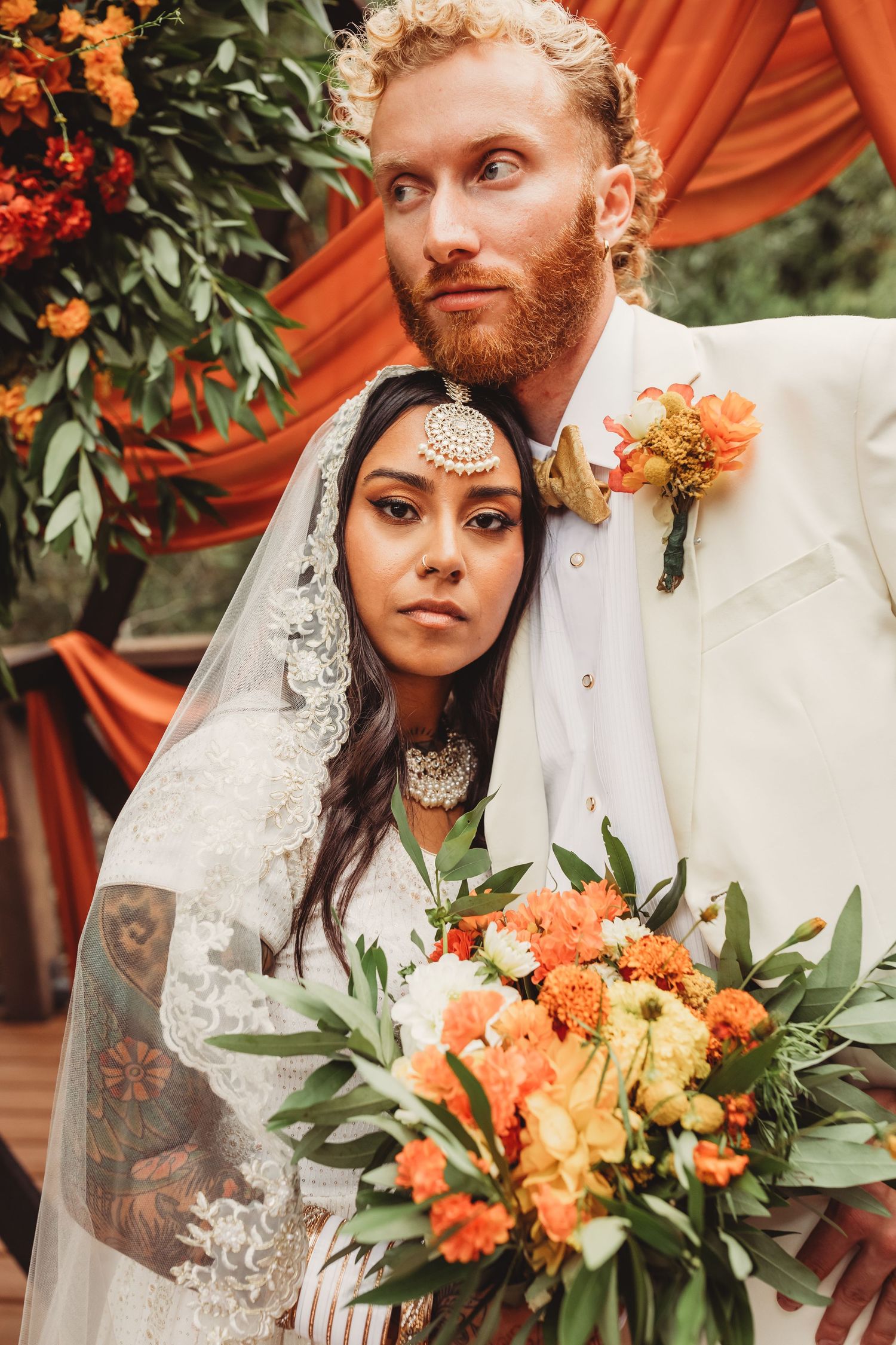 Romantic wedding couple poses against orange drapery with autumnal floral bouquet of peach and yellow blooms.