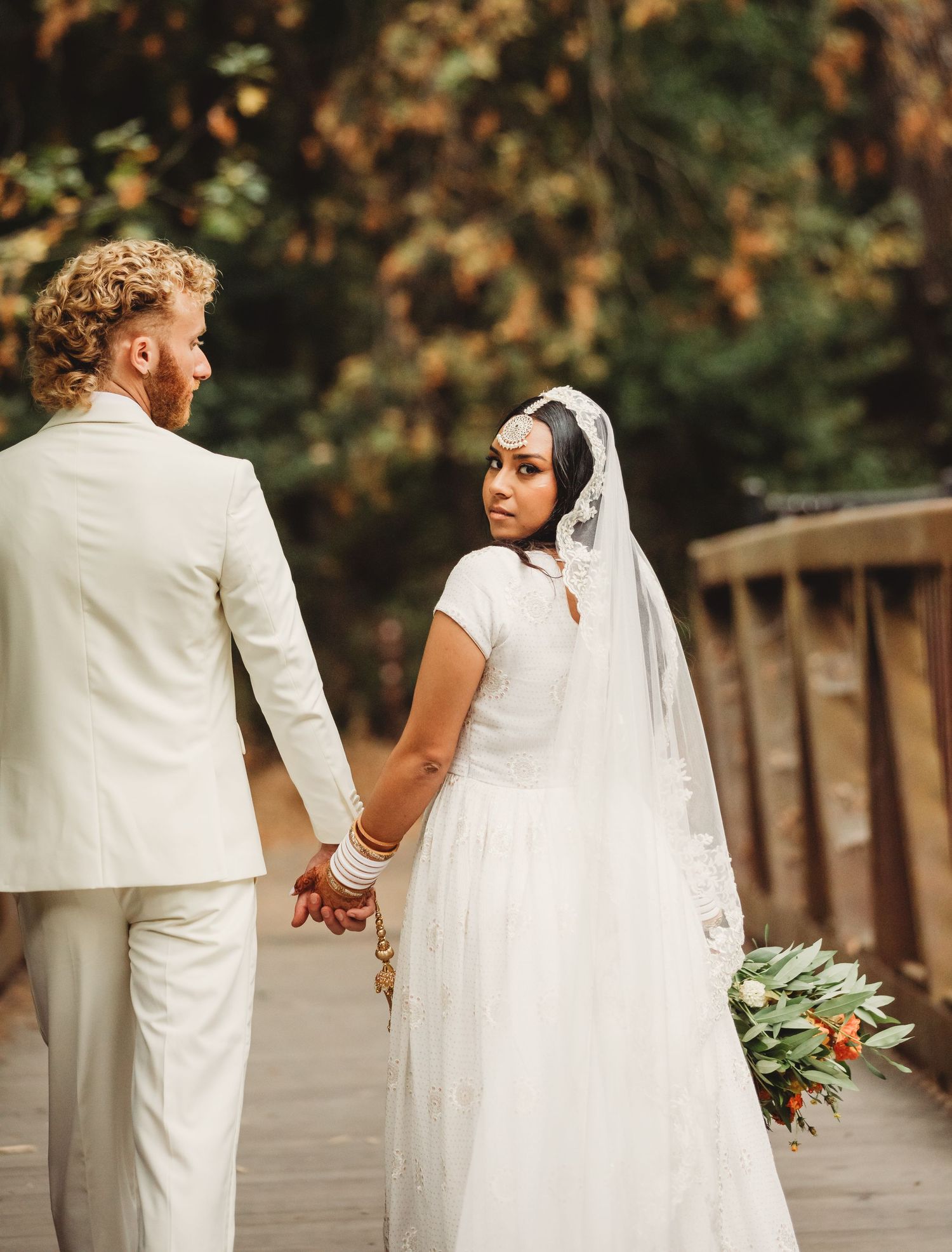 Couple in white wedding attire holding hands on wooden deck with autumn foliage in background.