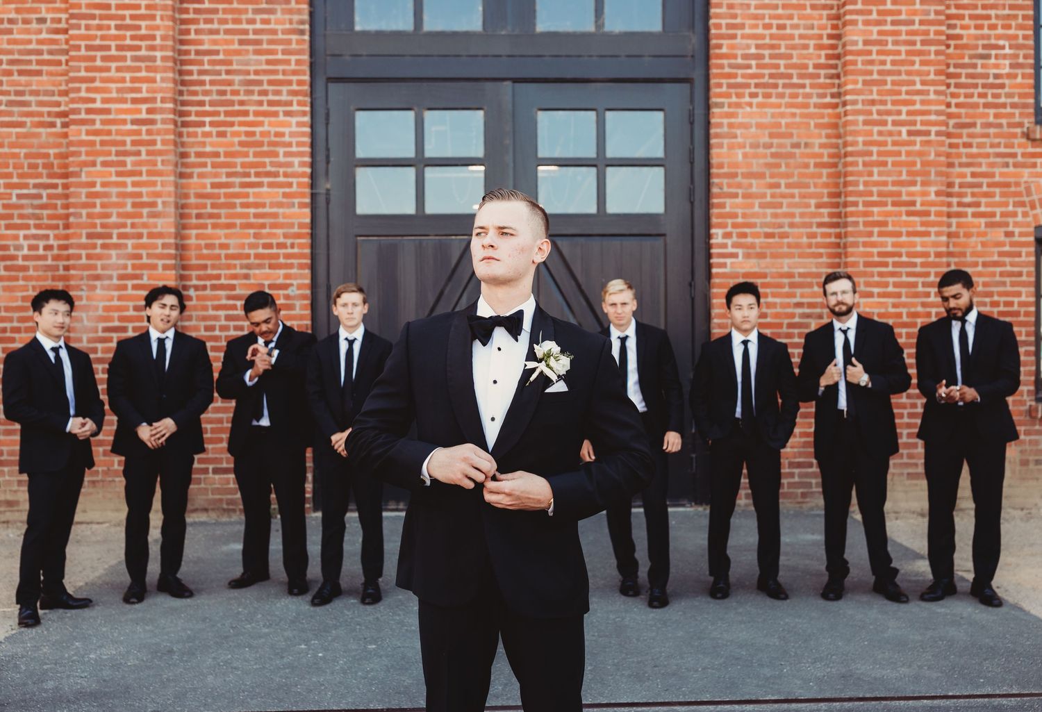 A groom in black tuxedo stands in front of groomsmen wearing dark suits against a red brick building.