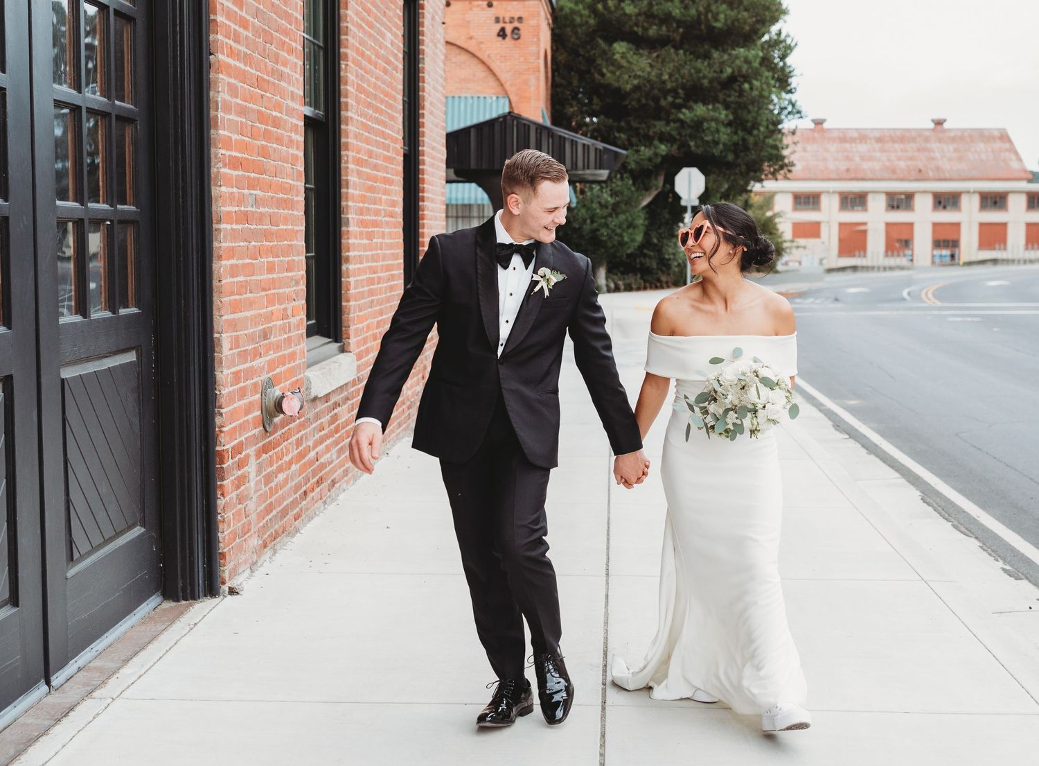 A bride and groom share intimate moments while walking along a brick-lined exterior wall of an urban building.