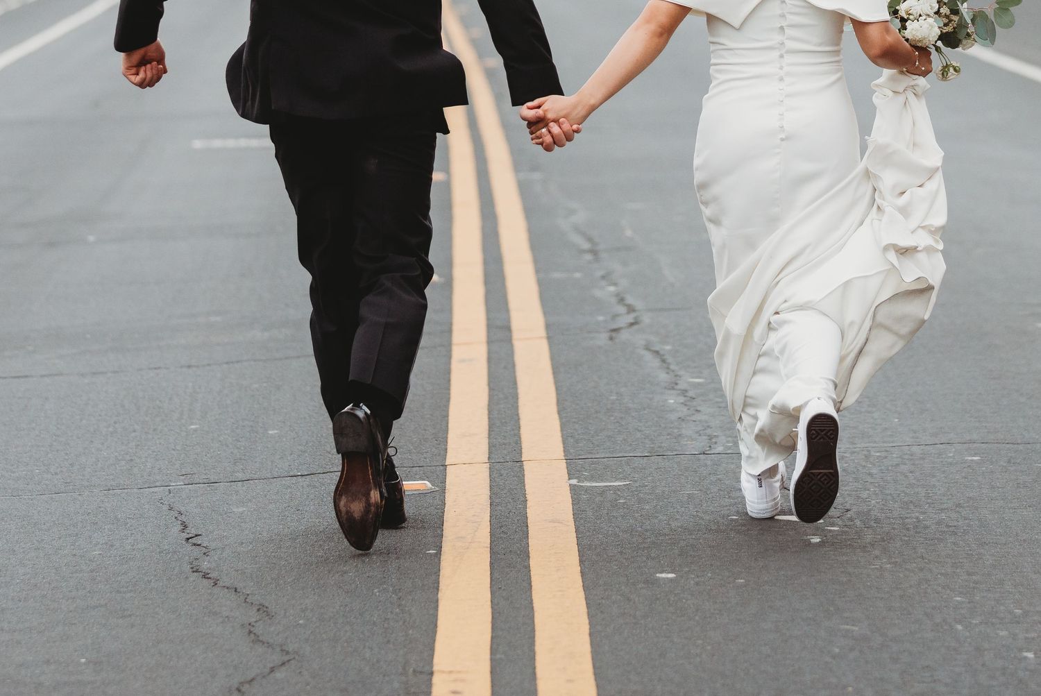 Bride and groom hold hands while walking along double yellow lines on a road, shown from waist down.
