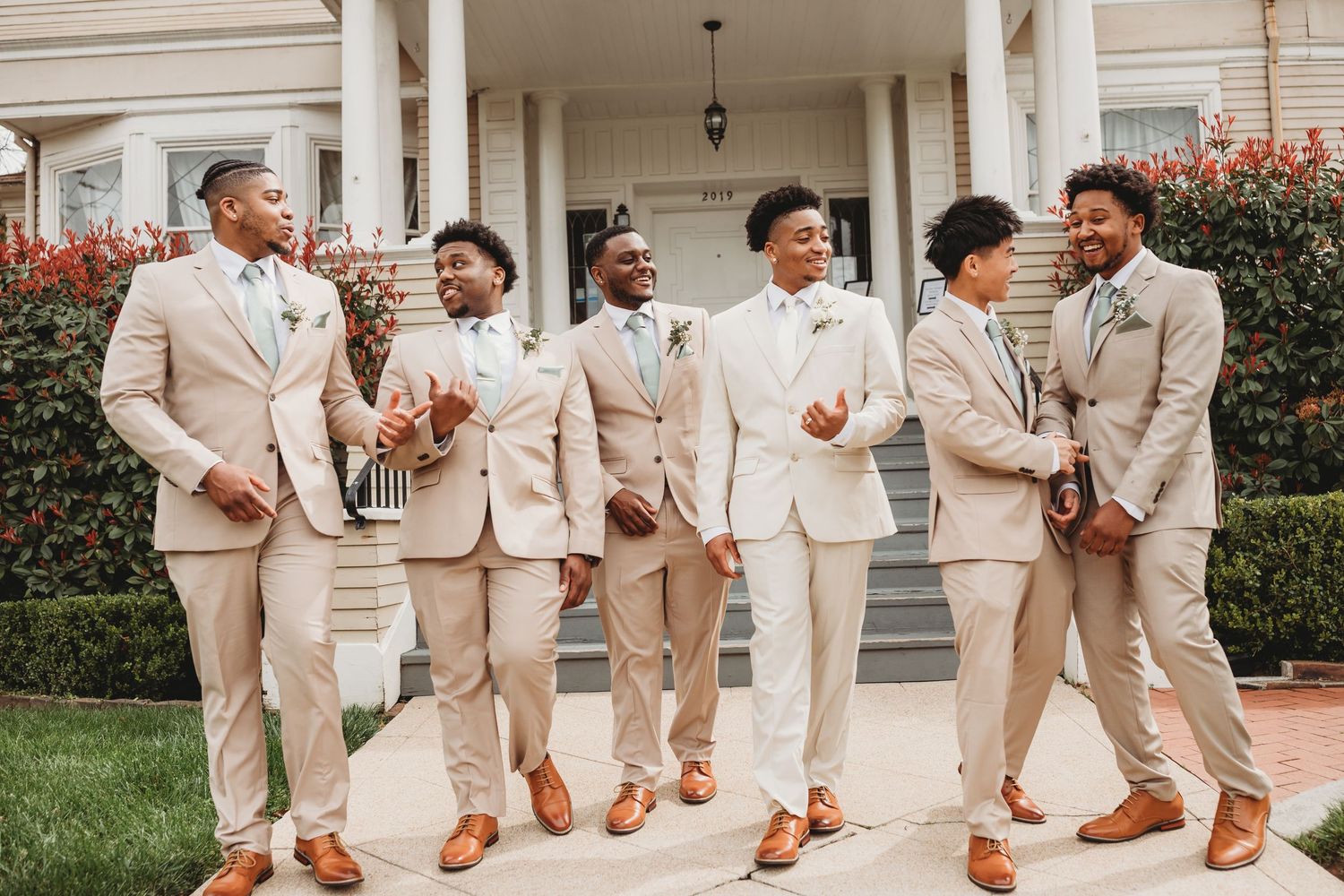 Groomsmen in beige suits strike fun poses on a columned porch.