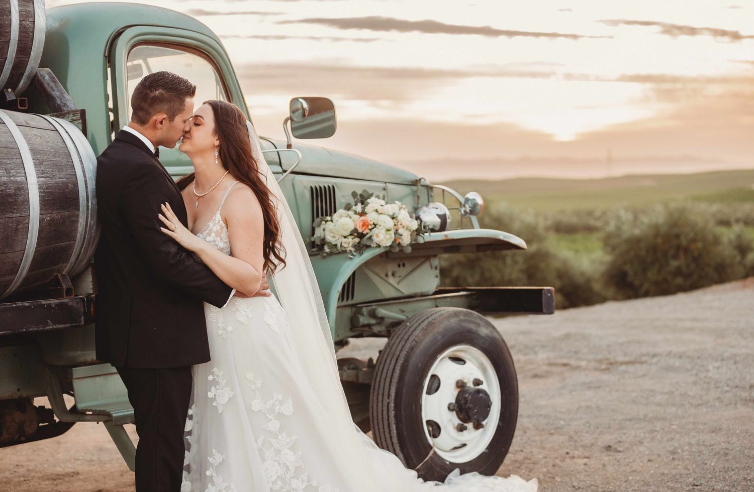A couple embraces beside a vintage green truck decorated with flowers during their rustic countryside wedding at sunset.