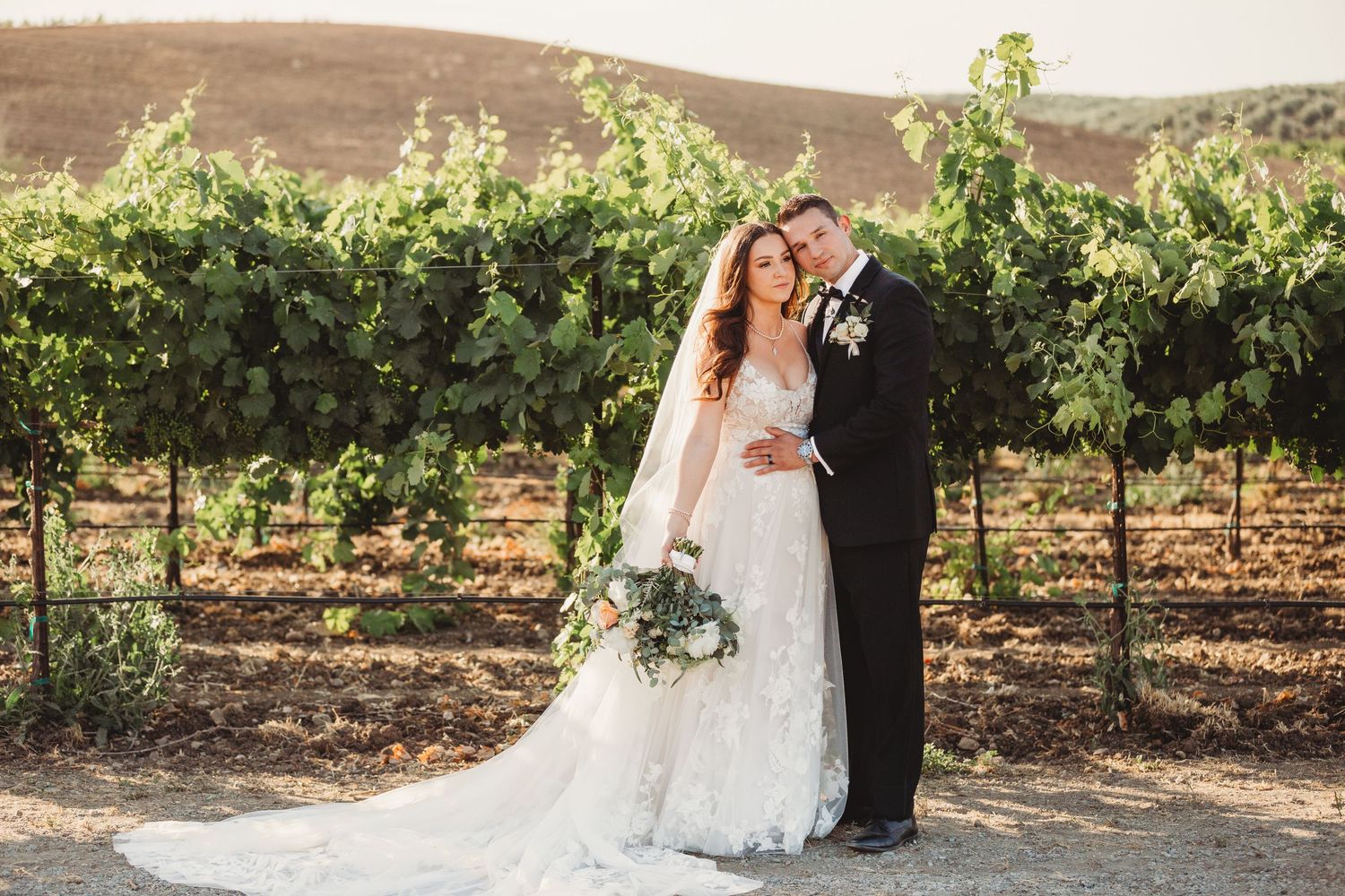 A romantic wedding portrait of a couple standing together in a scenic vineyard on a sunny day.