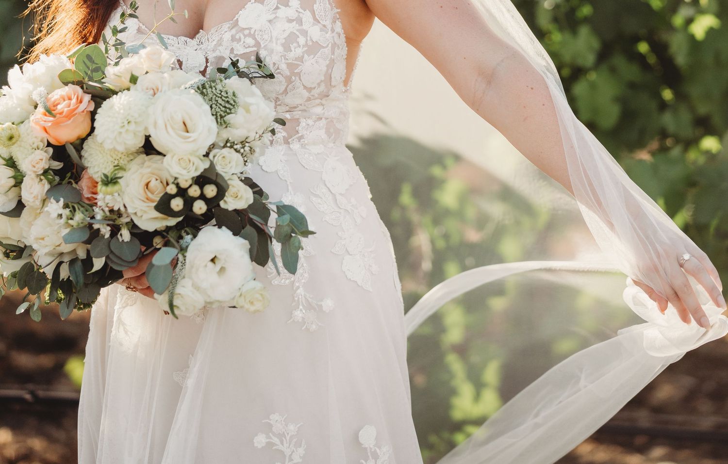 A bridal wedding dress with lace details flows gracefully in an outdoor garden setting with white floral bouquet.