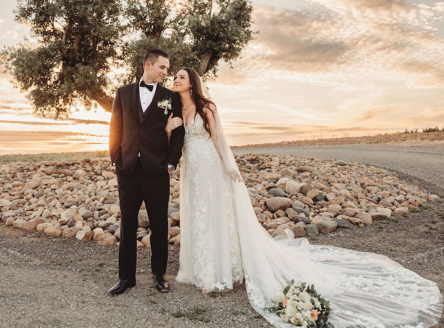 Couple shares elegant wedding photos at sunset in a desert landscape with trailing white dress and formal attire.